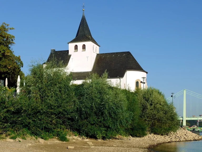 Foto der Kirche St. Nikolaus am Rheinufer in Köln-Rodenkirchen bei blauem Himmel, umgeben von Bäumen und der Rodenkirchener Brücke im Hintergrund.